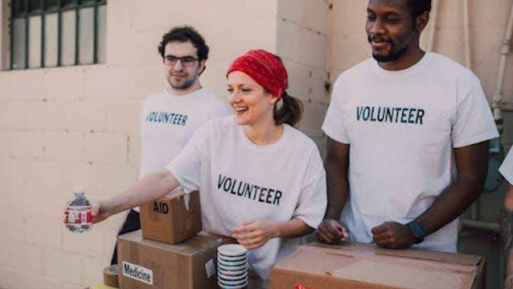 Three people in white “volunteer” shirts are handing out food and medicine.